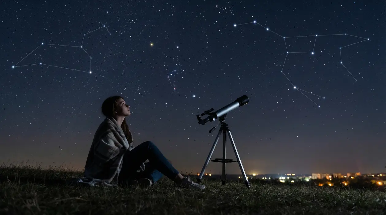 Una persona seduta su un prato guarda le stelle accanto a un telescopio sotto un cielo notturno stellato