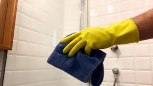 Una mano con guanto giallo pulisce il vetro della doccia con un panno blu